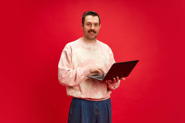 Man in sweatshirt standing with laptop isolated on red studio background. Remote work software demonstrating easy communication. Concept of human emotions, casual fashion, lifestyle, business - Photo, image
