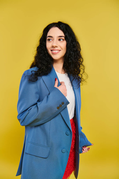 Mladá brunetka s kudrnatými vlasy udeří pózu ve stylové modré bundě, vyzařující sebevědomí a eleganci na žlutém pozadí. - Fotografie, Obrázek