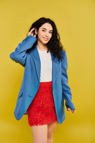 黄色の背景に自信とスタイルを除くスタイリッシュな青いジャケットと赤いスカートでカーリーヘアの若いブルネットの女性. - 写真・画像