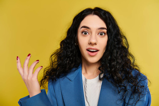 Een jonge brunette met krullend haar vertoont een verrassende expressie, toont haar emoties in een studio met een levendige gele achtergrond. - Foto, afbeelding