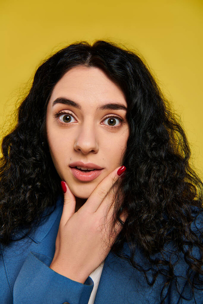 A spirited young woman with curly hair showcasing her emotions in a stylish blue jacket against a vibrant yellow background. - Photo, Image