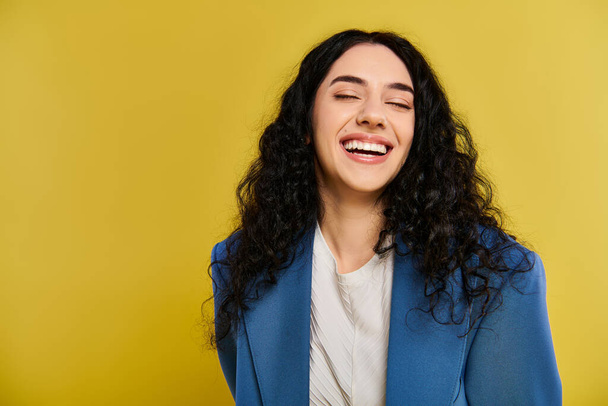 Молодая женщина с вьющимися волосами улыбается и носит голубую куртку, излучающую радость и уверенность в студийной обстановке с желтым фоном. - Фото, изображение