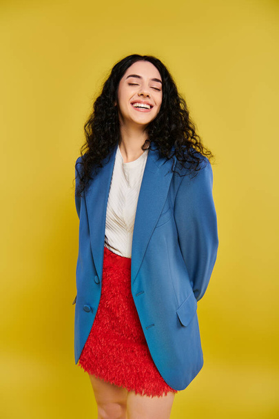 Young brunette woman with curly hair poses in stylish blue jacket and red skirt against yellow backdrop, showcasing her emotions. - Photo, Image