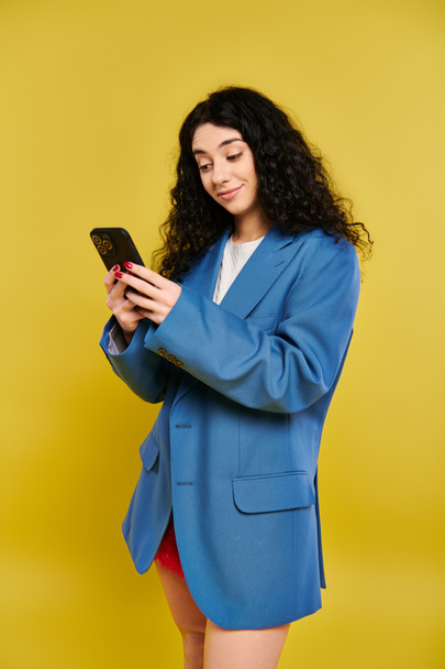 Μια νεαρή γυναίκα σε ένα μπλε σακάκι γοητευμένος από το κινητό της τηλέφωνο, έντονα εστιασμένη στην οθόνη. - Φωτογραφία, εικόνα