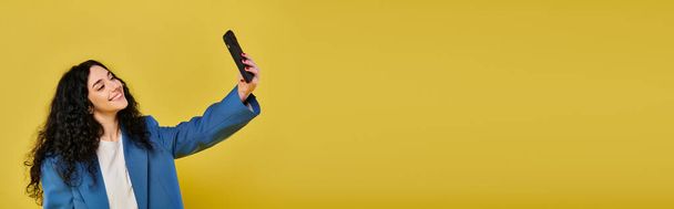 Eine brünette Frau in stylischer Kleidung posiert mit lockigem Haar und fängt ihre Emotionen mit einem Handy vor gelbem Hintergrund ein. - Foto, Bild