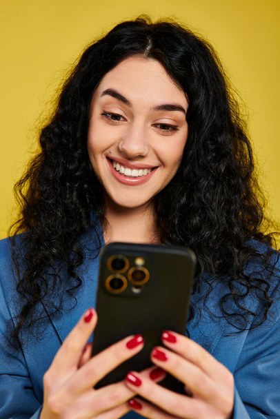 Una joven morena con el pelo rizado con un atuendo elegante expresando emociones mientras sostiene un teléfono celular, contra un fondo amarillo. - Foto, imagen