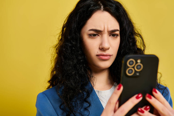 Egy fiatal barna nő göndör haj elegáns öltözékben tart egy mobiltelefon, megjelenítve a különböző érzelmek ellen sárga háttér. - Fotó, kép