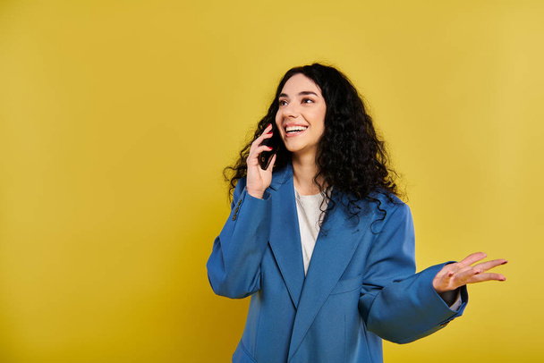 Jeune femme brune aux cheveux bouclés en veste bleue parlant sur téléphone portable dans une pose élégante sur fond jaune. - Photo, image