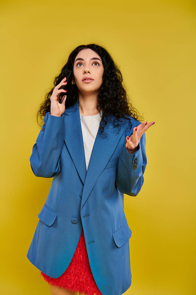 Brunette nainen kihara tukka poseeraa tyylikäs sininen bleiseri, esittelee tunteitaan studiossa keltainen tausta. - Valokuva, kuva
