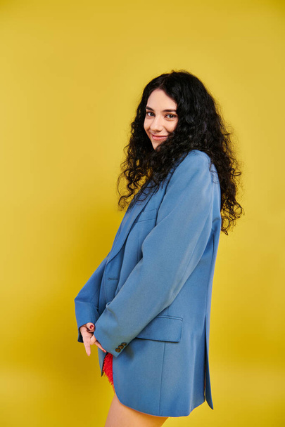 A young brunette woman with curly hair, in a blue suit, posing elegantly in a studio with a yellow background. - Zdjęcie, obraz