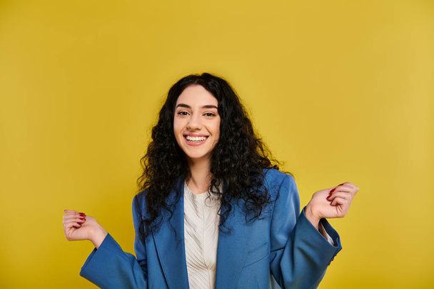 Молода жінка з кучерявим волоссям витончено позує в синій куртці і білій сорочці, демонструючи свої емоції на тлі жовтого студійного фону. - Фото, зображення
