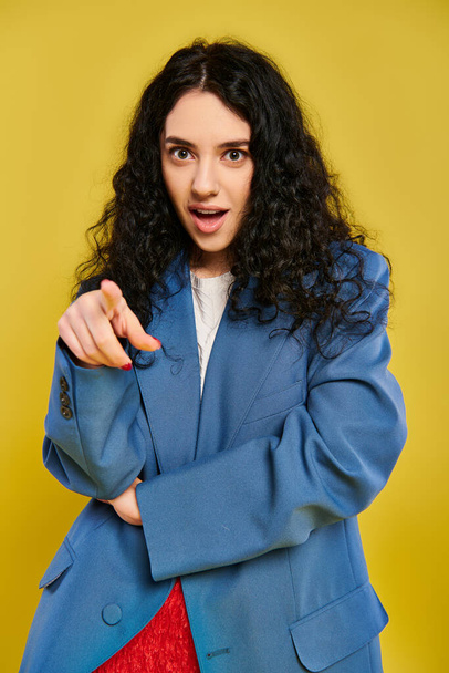 A young, brunette woman with curly hair striking a pose in a blue jacket while pointing directly at the camera. - Photo, Image