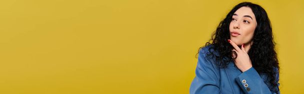 Young, curly-haired brunette woman poses confidently in a blue shirt in a studio with a vibrant yellow background. - Photo, Image