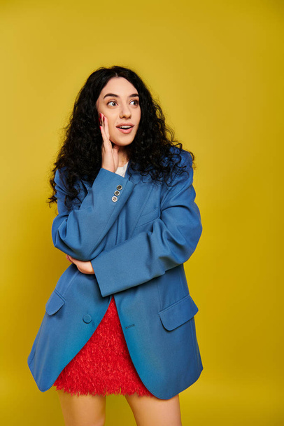 A young, brunette woman with curly hair poses for a picture in a stylish blue jacket, showcasing her emotions against a yellow background. - Photo, Image
