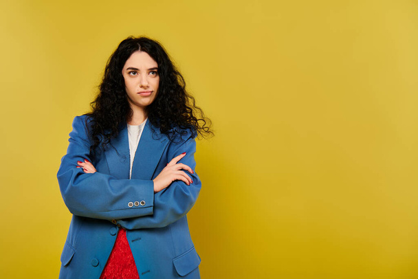A young brunette woman with curly hair confidently stands with her arms crossed in front of a vibrant yellow background. - Photo, Image