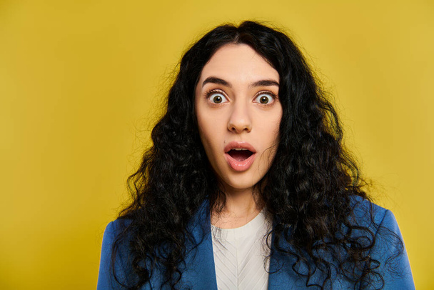 Μια νεαρή γυναίκα με σγουρά μαλλιά και κομψή ενδυμασία, με ανοιχτά μάτια και ανοιχτό στόμα, που εκφράζει γνήσια έκπληξη σε ένα στούντιο με κίτρινο φόντο. - Φωτογραφία, εικόνα