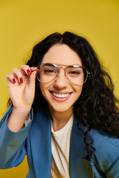 Giovane donna bruna con i capelli ricci posa con fiducia per un ritratto in uno studio, indossando occhiali eleganti su uno sfondo giallo. - Foto, immagini