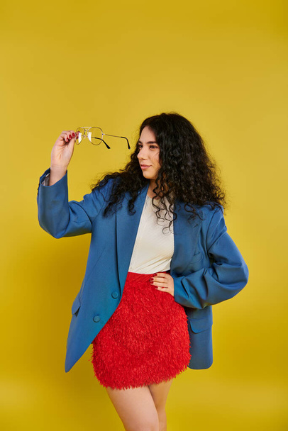 Una mujer joven y elegante con el pelo castaño rizado golpea una pose en una chaqueta azul vibrante y una falda roja llamativa contra un fondo amarillo soleado. - Foto, imagen