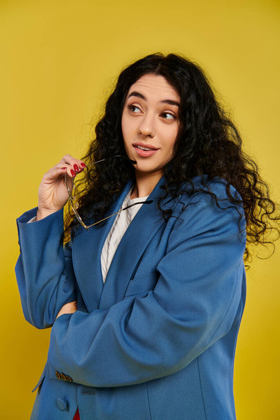 A young brunette woman with curly hair poses in a stylish blue jacket, showing glasses in a studio with a yellow background. - Photo, Image