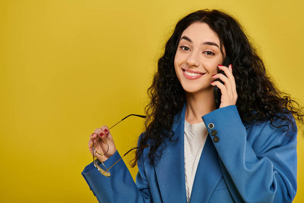 Een jonge brunette vrouw in een stijlvolle blauwe jas, praten op een mobiele telefoon met een gele achtergrond. - Foto, afbeelding