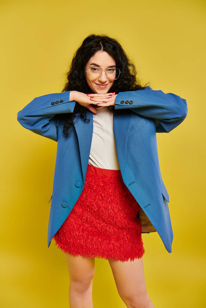 Une jeune femme brune aux cheveux bouclés pose dans une veste bleue élégante et une jupe rouge, mettant en valeur une gamme d'émotions dans un cadre de studio avec un fond jaune. - Photo, image