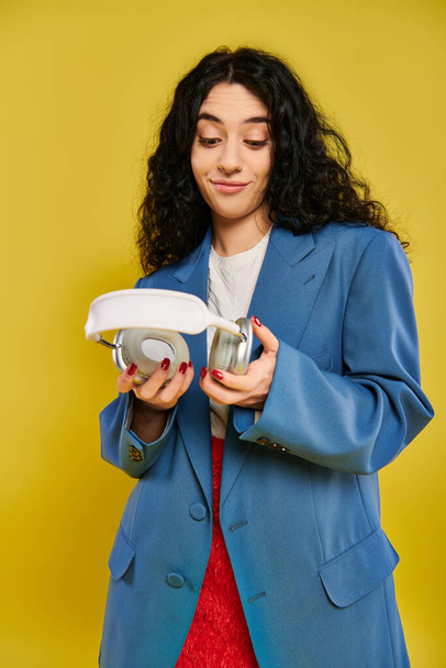 Eine junge brünette Frau mit lockigem Haar posiert in einem Studio-Setting und zeigt ihre Emotionen, während sie vor gelbem Hintergrund einen weißen Kopfhörer in der Hand hält.. - Foto, Bild