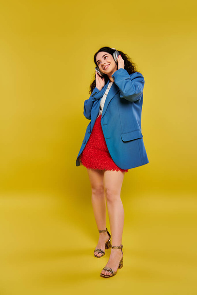 A young brunette woman with curly hair styled in a blue jacket and red dress using headphones - Photo, Image