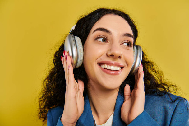 Een jonge brunette vrouw met krullend haar luistert naar muziek via de hoofdtelefoon, verloren in het ritme tegen een levendige gele achtergrond. - Foto, afbeelding