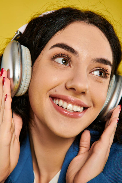 Una giovane donna elegante con i capelli ricci sorridente felicemente alla macchina fotografica mentre indossa le cuffie in uno studio con uno sfondo giallo. - Foto, immagini