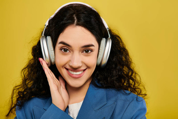Eine junge brünette Frau mit lockigem Haar und stilvoller Kleidung lächelt, während sie in einem Studio mit gelbem Hintergrund Kopfhörer trägt.. - Foto, Bild
