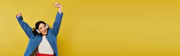 Giovane donna bruna con i capelli ricci in abbigliamento elegante, emotivamente alzando le braccia in uno studio giallo. - Foto, immagini