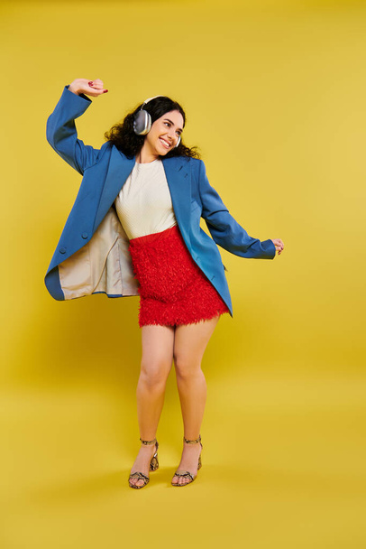A young brunette woman with curly hair emoting in a stylish blue jacket and red skirt against a bright yellow background. - Photo, Image
