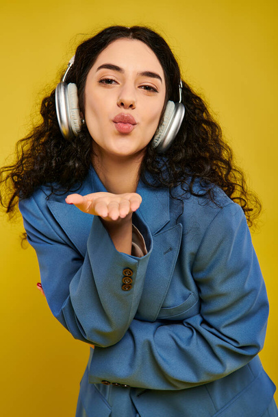Kıvırcık saçlı, genç, esmer bir kadın sarı arka planlı bir stüdyoda kulaklık takarken komik bir ifade takıyor.. - Fotoğraf, Görsel