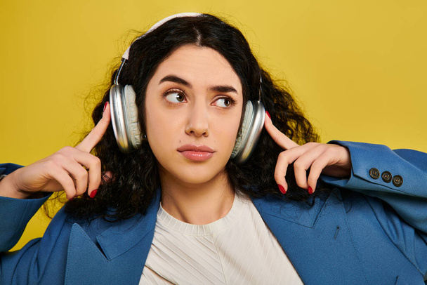 Stylová mladá žena s kudrnatými vlasy, se sluchátky, ponořená do hudby s klidným výrazem na žlutém pozadí. - Fotografie, Obrázek