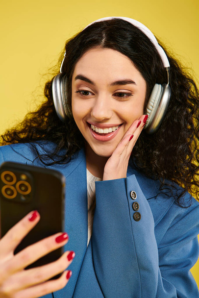 Młoda kobieta z kręconymi włosami, nosząca słuchawki i patrząca na komórkę, zanurzona w świecie muzyki i komunikacji. - Zdjęcie, obraz