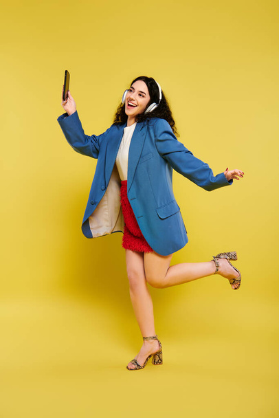 Young brunette woman with curly hair, dressed in a blue jacket and red skirt, holding a cell phone against a yellow backdrop. - Photo, Image
