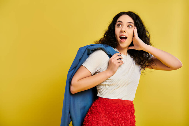 A young brunette woman with curly hair poses wearing a red skirt and blue jacket, showcasing her stylish and vibrant attire against a yellow studio background. - Photo, Image
