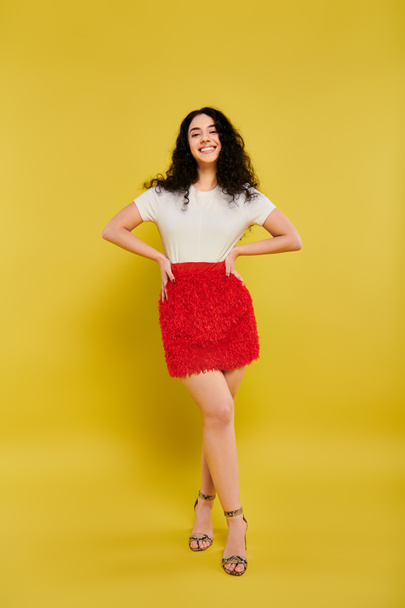 Brunette vrouw met krullend haar slaan een pose, presentatie van emoties in een stijlvolle rode rok tegen een gele achtergrond. - Foto, afbeelding