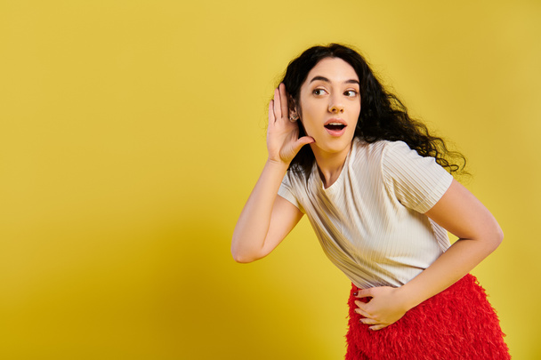 A brunette woman with curly hair looking surprised, dressed in stylish attire, in a studio with a yellow background. - Photo, Image