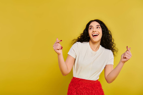 A young brunette woman with curly hair strikes a pose in a stylish red skirt and white shirt, showcasing her emotions against a yellow background. - Photo, Image
