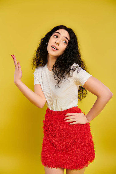 Uma jovem morena com cabelo encaracolado posa em uma saia vermelha impressionante, mostrando suas emoções em um estúdio com um fundo amarelo. - Foto, Imagem