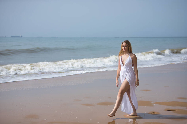 Молодая, красивая, блондинка в белом платье, гуляющая по одинокому пляжу, расслабленная и спокойная. Понятие мира, спокойствия, одиночества, обретения себя. - Фото, изображение