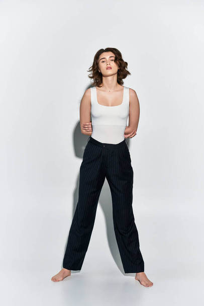 Ładna młoda kobieta w czarnych spodniach i biała bluzka ze zbiornika pewnie stoi pod czystą białą ścianą w studio.. - Zdjęcie, obraz