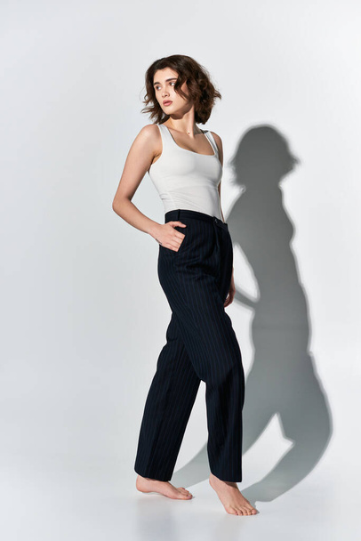 Een mooie jonge vrouw poseert vol vertrouwen in een zwarte broek en een wit topje, gracieus voor een witte muur. - Foto, afbeelding