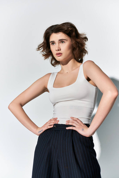 A stylish young woman strikes a strong pose, hands on hips, in black pants and white tank top, set against a grey backdrop. - Photo, Image