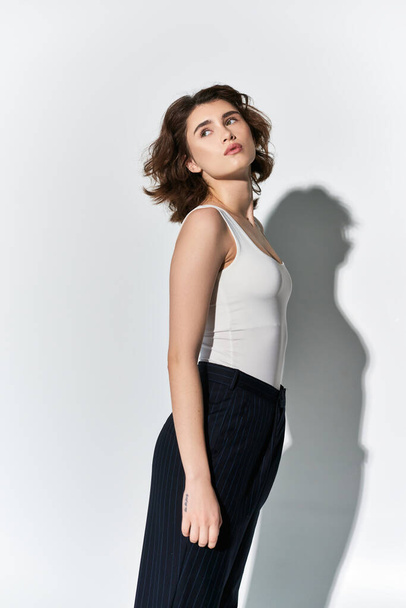 A pretty young woman in black pants and white tank top poses elegantly in front of a white wall in a studio setting. - Photo, Image