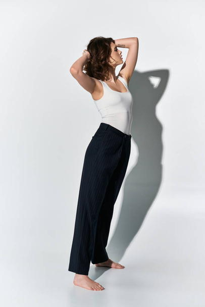 Uma jovem bonita posa em calças pretas e um top branco em um estúdio, de pé com confiança na frente de uma parede branca. - Foto, Imagem