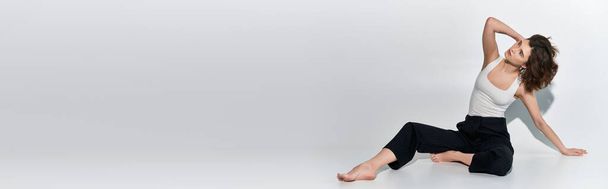 Спокойная молодая женщина в белой рубашке и черных штанах занимает позу в студии на сером фоне. - Фото, изображение