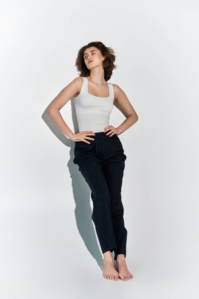 Een mooie jonge vrouw poseert sierlijk in een witte tank top en zwarte broek tegen een grijze studio achtergrond. - Foto, afbeelding