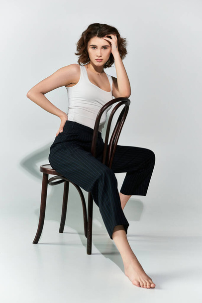 Śliczna młoda kobieta w czarnych spodniach i biała bluzka czołgowa z wdziękiem siedząca na drewnianym krześle w studio. - Zdjęcie, obraz